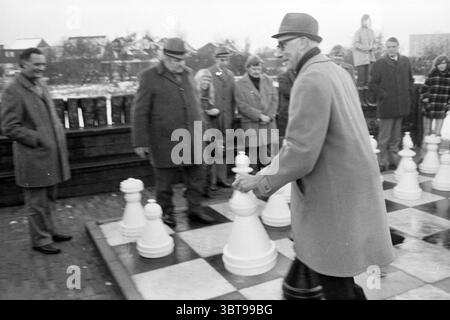 Eröffnung des Schachplatzes auf der Straße Beverwijk the Netherlands, Whizgle News, Dutch Desk, Niederlande, 1950 - 2000 am 30-11-1973. Diese Themen sind in der Abbildung dargestellt. Die Szene spielt in einem Außenbereich, wahrscheinlich in einem Park oder einem offenen platz, wo ein großes, übergroßes Schachbrett den Vordergrund dominiert. Die Schachfiguren sind massiv und bestehen aus weißem Material, das sich in starkem Kontrast zu den dunklen Quadraten des Bretts befindet, was einen Eindruck von einem strategischen Schlachtfeld vermittelt. Um das Schachbrett herum ist eine Gruppe von Menschen am Spiel beteiligt. Vorne steht ein Mann in einem hellen Mantel und einem Hut in m Stockfoto
