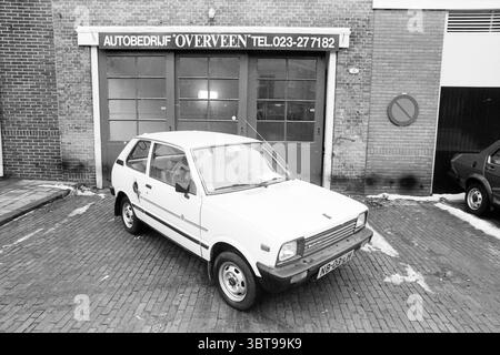 Anlass der Woche Overveen Overveen, Whizgle News, Dutch Desk, Niederlande, 1950 - 2000 am 17-02-1991. Diese Themen werden in der Abbildung angezeigt. Die Szene zeigt ein kompaktes, weißes Auto, das auf einer kopfsteingepflasterten Straße geparkt wird, dessen klare Linien sich in scharfem Kontrast zu den umgebenden Elementen bilden. Das Fahrzeug ist leicht außermittig positioniert, was die Aufmerksamkeit auf seine bescheidene Größe und das klassische Design lenkt. Hinter dem Auto bildet eine Ziegelwand den Hintergrund, die eine Mischung aus hellen und dunklen Ziegeltönen zeigt, die zu einem strukturierten Erscheinungsbild beitragen. Die Wand verfügt über große Garagentore, die in einem tiefen Schatten lackiert sind, was zusätzliche Details zur Folge hat Stockfoto