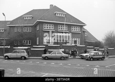 Erw. Oranje Nassauschool Z'voort Schools, Whizgle News, Dutch Desk, Niederlande, 1950 - 2000 am 19-12-1977. Die Abbildung zeigt diese Themen. Die Szene zeigt ein bescheidenes, zweistöckiges Backsteingebäude mit einem geneigten Dach, das sich durch symmetrische Fenster und klare weiße Zierleisten auszeichnet. Die Fassade aus dunkelroten Ziegeln trägt zu einem insgesamt soliden Erscheinungsbild bei. Um die Struktur herum versammelt sich eine Gruppe von Menschen – hauptsächlich Kinder mit wenigen Erwachsenen – in einer animierten Art und Weise, was eine lebhafte Atmosphäre suggeriert. Die Kinder sind in einer Mischung aus lebhaften Farben gekleidet, die dem M einen verspielten Kontrast verleihen Stockfoto