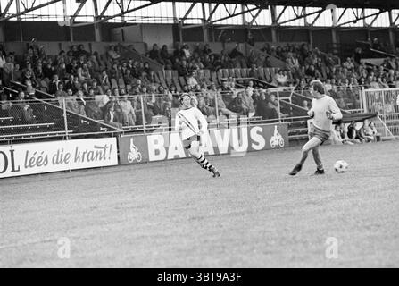 Final Velser Amateur Football (Velsen - IJmuiden) Football V.S.V. Velsen Stormvogels, Whizgle News, Dutch Desk, Niederlande, 1950 - 2000 am 11-05-1976. Das Bild enthält diese Themen. Die Szene zeigt ein lebhaftes Fußballspiel auf einem gut gepflegten Grasfeld, umgeben von einem stabilen Metallzaun. Auf der einen Seite läuft ein Spieler in einer hellen Uniform, möglicherweise weiß, energisch auf den Ball zu, der auf dem Spielfeld rollt. Seine Uniform hat markante schwarz-weiße Streifen, die zu einem auffälligen Kontrast zum leuchtenden grünen Gras beitragen. Im Hintergrund ist ein Torhüter, c Stockfoto