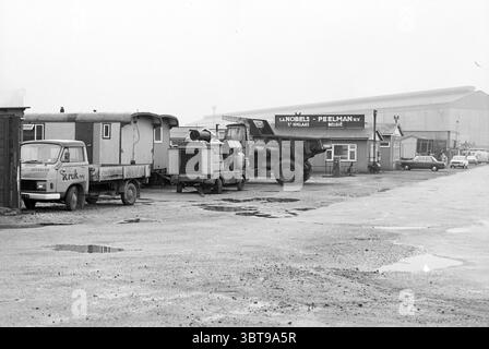 Business Park Haarlem, Niederlande, Whizgle News, Dutch Desk, Niederlande, 1950 - 2000. Das Bild enthält diese Themen. Die Szene zeigt ein etwas industrielles Gebiet, das durch eine Mischung von Fahrzeugen und Strukturen gekennzeichnet ist. Im Vordergrund befinden sich mehrere Fahrzeuge auf einer gepflasterten Oberfläche, die Anzeichen von Verschleiß zeigt, mit mehreren großen Pfützen, die den bewölkten Himmel reflektieren. Auf der linken Seite steht ein kleiner, klassischer Lkw in Kastenform mit gedämpfter Farbe. Daneben steht ein etwas größeres Fahrzeug, möglicherweise ein Tieflader oder ein Nutzfahrzeug, auf dem geparkt wird. Beide Lkws erscheinen veraltet, was ein Gefühl o hervorruft Stockfoto