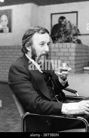 Mann mit Cognacglas im modernen Gemeindezentrum., Whizgle News, Dutch Desk, Niederlande, 1950 - 2000. Dies sind die Themen im Bild. Die Szene zeigt einen Gentleman, der bequem in einem modernen, minimalistischen Stuhl sitzt und einen Hauch von Raffinesse ausstrahlt. Er hat einen gepflegten Bart und ein leicht welliges Haar, was auf sein reifes Alter hindeutet. Seine Kleidung ist scharf und zeichnet sich durch einen dunklen Nadelstreifenanzug aus, der ein Gefühl von Professionalität und Eleganz vermittelt. Der Anzug wird durch ein helles Hemd ergänzt, das am Kragen sichtbar ist. In einer Hand hält er ein Glas, das eine dunkle Flüssigkeit enthält, möglicherweise ein Whir Stockfoto