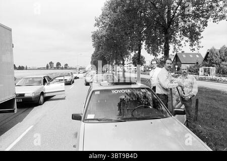 Die Autos auf der Straße hielten wegen Staus an, die Fahrer stiegen aus, Whizgle News, Dutch Desk, Niederlande, 1950 - 2000 am 07-1989. Das Bild enthält diese Themen. Die Szene zeigt eine Situation am Straßenrand mit mehreren Autos, die entlang einer von Bäumen gesäumten Allee geparkt sind. Bei den Fahrzeugen handelt es sich meist um klassische Modelle mit einer Vielzahl von Farben, darunter Grau-, Blau- und Weißtöne. Ein paar Türen sind offen, was darauf hinweist, dass sich die Passagiere entweder drinnen befinden oder gerade aus dem Hotel getreten sind. Zwei Männer stehen in der Nähe des Vordergrunds und unterhalten sich. Einer scheint sich beiläufig an ein Auto zu lehnen, während der andere gestikuliert Stockfoto