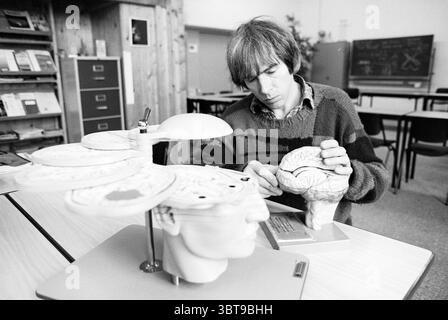 Mr. Rob Raam Coma Patient, Whizgle News, Dutch Desk, Niederlande, 1950 - 2000 auf 31-01-1994. Das Bild enthält diese Themen. Die Szene findet in einem Klassenzimmer oder einem Arbeitsbereich statt, der sich durch ein warmes, hölzernes Interieur mit hellen Holztäfelungen an den Wänden auszeichnet. Die Atmosphäre vermittelt eine gelehrte und besinnliche Stimmung, die durch sanfte, diffuse Umgebungsbeleuchtung verstärkt wird. Im Vordergrund konzentriert sich eine Person, ein Mann mit schulterlangen Haaren, darauf, ein Modell des Gehirns zusammenzustellen. Er trägt einen gestreiften Pullover in Grün- und Brauntönen, der seinem Erscheinungsbild einen lässigen Touch verleiht Stockfoto