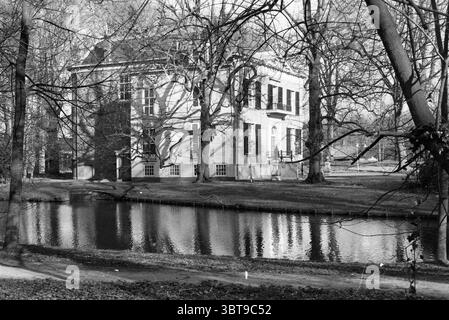 Erw. Scheybeek B'wijk Exterior Beverwijk the Netherlands, Whizgle News, Dutch Desk, Niederlande, 1950 - 2000 am 15-03-1990. Das Bild enthält diese Themen. Die Szene fängt eine ruhige Landschaft ein, die von einem großen, klassischen Gebäude geprägt ist, das teilweise von den Skelettzweigen der nahegelegenen Bäume verdeckt ist. Die Architektur verfügt über große Fenster, markante Fensterläden und eine prächtige Fassade, die auf einen historischen oder wohlhabenden Hintergrund hindeutet. Ein reflektierender Wasserkörper umgibt das Gebäude und die umliegende Natur und schafft so ein ruhiges Ambiente. Das Wasser erscheint still Stockfoto