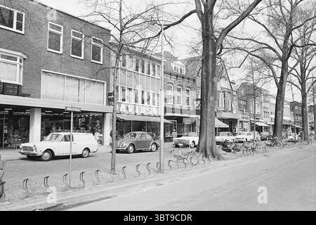 Street View Shops in Breestraat nahe Davidson Herrenmode Beverwijk Breestraat Niederlande, Whizgle News, Dutch Desk, Niederlande, 1950 - 2000 am 01-04-1969. Das Bild enthält diese Themen. Die Szene fängt eine lebhafte Stadtstraße mit einer Reihe von Ladenfronten ein, die jeweils unterschiedliche architektonische Stile aufweisen, die eine Mischung aus modernen und klassischen Einflüssen suggerieren. Die Gebäude weisen große Glasfenster auf, die ein Gefühl von Offenheit schaffen, während ihre Fassaden durch Ziegelsteine und Mauerwerk gekennzeichnet sind, die verschiedene Grau- und Brauntöne zeigen. An der Bordsteinkante stehen mehrere Oldtimer, Stockfoto