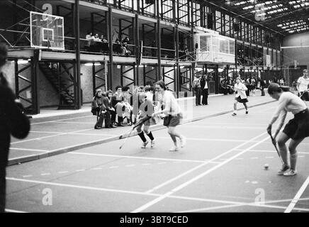 Finales Hockeyturnier Saxenburgers (Bennebroek - Cartouche) Hockey, Whizgle News, Dutch Desk, Niederlande, 1950 - 2000 am 26.12.1977. Die Abbildung zeigt diese Themen. Die Szene zeigt eine belebte Halle mit einem großen Platz, der für ein Feldhockeyspiel organisiert wird. Auf der einen Seite stehen zwei Spieler in einem intensiven Moment, wobei einer dunkle Sportausrüstung und der andere ein leichteres Outfit trägt, beide konzentrieren sich auf die Ballkontrolle. Ihre Körper befinden sich in dynamischer Bewegung, was auf die Wettbewerbscharakter des Spiels hinweist. Um den Platz herum füllen die Zuschauer die Tribünen und sind Meer Stockfoto
