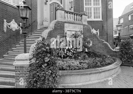 Blumenbeet vor dem Rathaus Z'voort Zandvoort, Whizgle News, Dutch Desk, Niederlande, 1950 - 2000 am 07-06-1995. Das Bild enthält diese Themen. Die Szene fängt eine architektonische Umgebung mit komplizierten Details und einem Sinn für Geschichte ein. Eine große Treppe, flankiert von kunstvollen Geländern, führt zu einem wunderschön gestalteten Eingang. Die Stufen bestehen aus Stein, deren Oberfläche Anzeichen von Verschleiß aufweist, was auf einen langen Lauf der Zeit hindeutet. Vor dem Treppenhaus fällt üppiges Grün über ein rundes Blumenbeet, das von einer Vielzahl von Pflanzen in voller Blüte überschwemmt wird. Farbtöne von wh Stockfoto