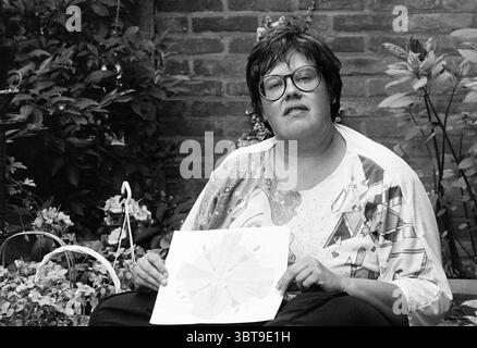 Trees Klaasen H'dorp Hoofddorp the Netherlands, Whizgle News, Dutch Desk, Niederlande, 1950 - 2000 am 02-07-1992. Dies sind die Themen im Bild. In einer ruhigen Umgebung im Freien sitzt eine Person bequem zwischen lebendigen Blumen und Grün. Der Hintergrund besteht aus rustikalen Ziegeln, die der Szene einen warmen, erdigen Ton verleihen. Die Person hat kurze, lockige Haare und trägt eine Brille, die ein freundliches Auftreten zeigt. Ihre Kleidung hat ein farbenfrohes, abstraktes Design, das einen schönen Kontrast zur natürlichen Umgebung bildet und auf eine kreative Persönlichkeit hinweist. In ihren Händen halten sie ein dekoratives A Stockfoto