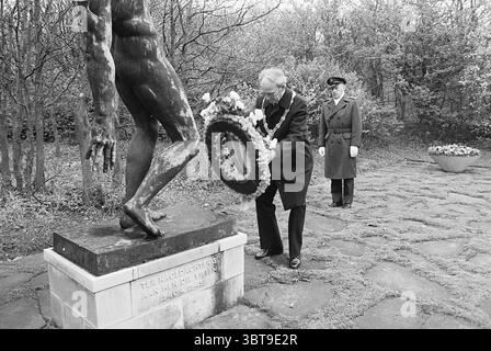 Kranzlagen auf dem Friedhof Duinhof Gedenktag Kranzlagen, Whizgle News, Dutch Desk, Niederlande, 1950 - 2000 am 04-05-1977. Diese Themen sind in der Abbildung dargestellt. In einer düsteren Umgebung im Freien sind zwei Figuren der Mittelpunkt vor einem Hintergrund von Bäumen, die ein schwach verschwommenes grünes Baldachin bilden. Die Szene ist in Grautönen gerendert, was eine melancholische Atmosphäre verleiht. Eine Person, die in formeller dunkler Kleidung gekleidet ist, kniet vor einer auffälligen Statue, die eine muskulöse Figur mit ausgestreckten Armen darstellt, die Stärke und vielleicht Widerstandsfähigkeit symbolisiert. Die Oberfläche der Statue wirkt strukturiert und stoi Stockfoto