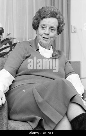 Frau Heetveld Kindergarten Ausbildung Inspector Personal Education, Whizgle News, Dutch Desk, Niederlande, 1950 - 2000 am 22-12-1977. Diese Themen werden in der Abbildung angezeigt. Die Szene zeigt eine ältere Frau, die bequem in einem Plüschstuhl sitzt. Sie hat einen warmen Ausdruck, mit sanft geschwungenen Lippen in einem subtilen Lächeln. Ihr Haar ist mit weichen, voluminösen Locken gestylt und zeigt einen klassischen Look. Sie trägt ein bescheidenes, aber stilvolles Outfit, das sich durch ein kurzärmeliges Oberteil über einem hochausgeschnittenen Kleidungsstück und einen knielangen Rock auszeichnet, der bequem zu ihr passt. Die Farbpalette bietet gedämpfte Töne; die Kleidung i Stockfoto