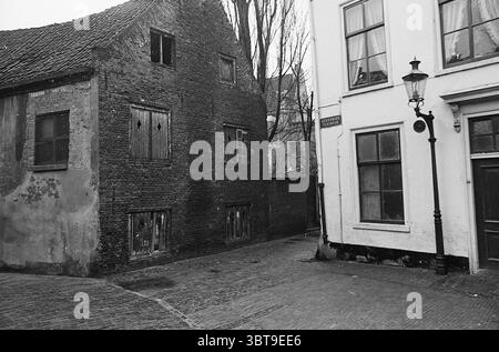 Gesicht von Haarlem Haarlem Goudsmidspleintje The Netherlands, Whizgle News, Dutch Desk, Niederlande, 1950 - 2000 am 23-03-1965. Dies sind die Elemente im Bild. Die Szene fängt eine krasse, urbane Umgebung mit einem unverwechselbaren, historischen Charakter ein. Zwei Gebäude flankieren eine enge Kopfsteinpflasterstraße und schaffen ein Gefühl von Intimität. Auf der einen Seite befindet sich eine alte Ziegelstruktur, die durch dunkle, verwitterte Oberflächen gekennzeichnet ist. Die Fenster sind verschachtelt, was auf Vernachlässigung oder Vernachlässigung hindeutet; einige Holzlatten schälen oder fehlen vollständig, was die baufällige Atmosphäre verstärkt. Die Textur des Ziegels ist rau, mit Stockfoto