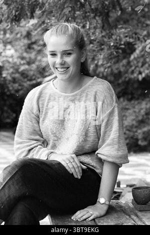 Leonie Voelink ist ein Filmstar, der Filmaufnahmen filmt, Whizgle News, Dutch Desk, Niederlande, 1950 - 2000 am 07-09-1986. Dies sind die Themen im Bild. In einer ruhigen Umgebung im Freien sitzt eine junge Frau bequem auf einer rustikalen Holzbank. Sie hat ein helles Lächeln, das Wärme und Zugänglichkeit ausstrahlt. Ihr heller, übergroßer Strickpullover bildet einen sanften Kontrast zu ihrer dunklen Jeans und sorgt so für einen lässigen, aber schicken Look. Eine üppige grüne Kulisse um sie herum verleiht ihr einen lebendigen Touch, mit verschiedenen Grüntönen der Blätter und Äste, die sie umrahmen. Die Atmosphäre fühlt sich entspannt an Stockfoto