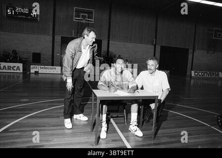 Unterzeichnen Sie einen Vertrag mit dem Basketball Club, Whizgle News, Dutch Desk, Niederlande, 1950 - 2000 am 27.04.1990. Diese Themen werden in der Abbildung angezeigt. In einem geräumigen Indoor-Sportsaal stehen zwei Männer neben einer sitzenden Person an einem kleinen Tisch. Die Umgebung verfügt über Holzfußböden, die charakteristisch für einen Fitnessraum sind, mit hellen Wänden und einer hohen Decke. Das Licht ist hell, wahrscheinlich kommt es von Deckenleuchten, wodurch eine gut beleuchtete Szene entsteht, die die Aktivität am Tisch betont. Der sitzende Mann trägt sportliche Freizeitkleidung, einschließlich einer locker sitzenden Jacke, die ein lebendiges Design zeigt Stockfoto