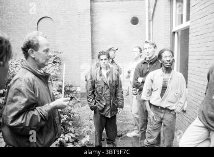 Marinekrankenhaus besetzte, Whizgle News, Dutch Desk, Niederlande, 1950 - 2000 am 14-09-1991. Das Bild enthält diese Themen. Die Szene zeigt eine Gruppe von Einzelpersonen, die in einem engen Raum stehen, der wie ein Innenhof oder Durchgang zwischen Gebäuden aussieht. Die Wände sind aus rotem Backstein gefertigt und schaffen eine warme, aber gedämpfte Kulisse. Im Vordergrund spricht ein Mann mit kurzen, lockigen Haaren mit den Händen, gekleidet in einer dunklen, strukturierten Jacke. Er hat einen engagierten Ausdruck, der möglicherweise eine wichtige Botschaft oder Anweisungen vermittelt. Um ihn herum sind mehrere Menschen unterschiedlichen Alters und Stils Stockfoto