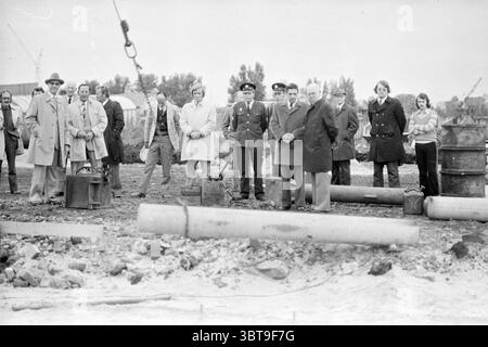 Fahrt des ersten Stapels für ein Bauprojekt, Whizgle News, Dutch Desk, Niederlande, 1950 - 2000 im Jahr 1975. Dies sind die Themen im Bild. Die Szene zeigt eine Gruppe von Individuen, die sich im Freien versammelt haben, wahrscheinlich an einem Bau- oder bahnbrechenden Ort. Der Hintergrund zeigt eine karge Industrielandschaft mit wenigen Strukturen wie Lagerhäusern oder Fabriken, die bei bewölktem Himmel leicht sichtbar sind. Im Vordergrund stehen verschiedene Männer und Frauen in etwas formeller Kleidung, was darauf hindeutet, dass sie für einen wichtigen Anlass da sind. Die Männer sind in Mänteln und Anzügen gekleidet, mit Sever Stockfoto