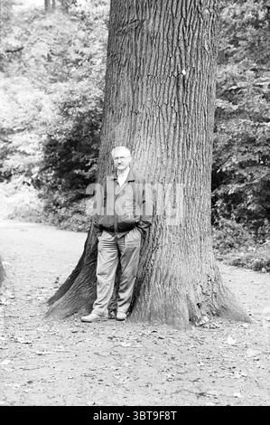 Herr Duinker Baumexperte de Hout Trees, Whizgle News, Dutch Desk, Niederlande, 1950 - 2000 am 19.09.1989. Die Abbildung zeigt diese Themen. In einer ruhigen, waldreichen Umgebung steht ein Mann neben einem massiven, strukturierten Baum, dessen Rinde tiefe Rillen und Grate aufweist und der Landschaft Charakter verleiht. Der große Stamm des Baumes dominiert den Vordergrund und betont sein Alter und seine Festigkeit. Der Mann, der in einer dunklen Jacke und einer hellen Hose gekleidet ist, lehnt sich lässig an den Baum, mit einer entspannten Haltung, die Komfort in dieser natürlichen Umgebung suggeriert. Um ihn herum gibt es verschiedene Grüntöne aus den Höhlen Stockfoto