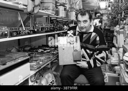 Guus Hermans Wassersportgeschäft Kennemerlaan IJmuiden schlägt IJmuiden Kennemerlaan Niederlande, Whizgle News, Dutch Desk, Niederlande, 1950 - 2000 am 15-02-1989. Diese Themen sind in der Abbildung dargestellt. In einem übersichtlichen Innenbereich sitzt ein Mann im Vordergrund. Er trägt einen dicken, gemusterten Pullover mit auffälligen geometrischen Formen in Schwarz, weiß und Grau, der der ansonsten dezenten Farbpalette ein Gefühl von Wärme verleiht. Sein Ausdruck deutet auf eine Mischung aus Stolz und Nostalgie hin, während er ein Holzbrett mit vielen Texturen hochhält, das mit verschiedenen Gegenständen verziert ist, die auf ein künstlerisches o hindeuten Stockfoto