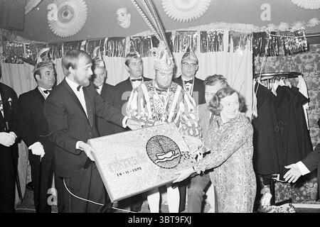 Tabelle der Verdienste Zandvoort Award Ceremony, Whizgle News, Dutch Desk, Niederlande, 1950 - 2000 am 14-11-1970. Das Bild enthält diese Themen. In einem festlichen Innenbereich versammelt sich eine Gruppe gut gekleideter Personen um eine zentrale Figur, die in aufwendiger Kleidung geschmückt ist, was auf einen feierlichen Anlass hinweist. Im Mittelpunkt der Szene steht ein Mann in einem auffälligen Kostüm, das mit komplizierten Mustern und Texturen verziert ist und einen Kopfschmuck trägt, der sein königliches Aussehen unterstreicht. Er steht selbstbewusst in der Mitte und hält eine große, reich verzierte Kiste, die ein Geschenk zu sein scheint. Er flankiert von einer Frau, die sich verkleidet Stockfoto