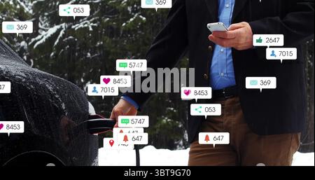 Blazer-verkleideter Mann, der das Kabel in den Autohafen in verschneiten Wäldern einsteckt, mit Smartphone und Benachrichtigungssymbolen Stockfoto