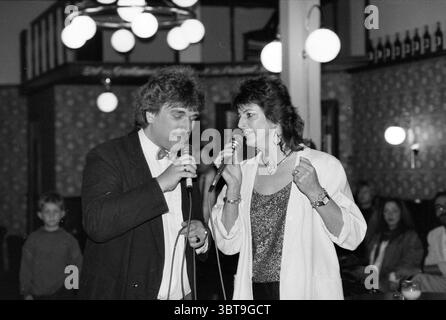 Haarlems Zijlpoort Gesangsfestival Singen und Chöre Haarlem the Netherlands, Whizgle News, Dutch Desk, Niederlande, 1950 - 2000 am 16.04.1989. Das Bild enthält diese Themen. Die Szene spielt in einem lebhaften Innenraum, wahrscheinlich in einer Bar oder einem kleinen Bühnenbereich, der von einer warmen und einladenden Atmosphäre geprägt ist. Die Wände sind mit gemusterten Tapeten verziert, die eine nostalgische Kulisse schaffen. Im Vordergrund stehen ein Mann und eine Frau in einem Duett, die jeweils ein Mikrofon halten. Der Mann, gekleidet in einem klassischen schwarzen Anzug mit einem weißen Hemd und einer Fliege, hat eine selbstbewusste Haltung, während er singt. Sein Haarstyl Stockfoto