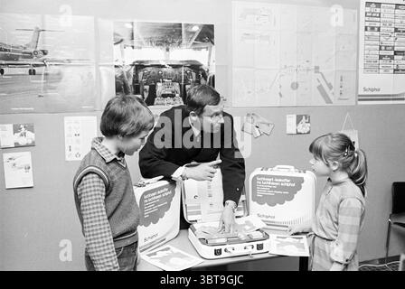 Präsentation des Luftfahrtlehrkoffer Schiphol Schiphol, Whizgle News, Dutch Desk, Niederlande, 1950 - 2000 am 23-04-1980. Diese Themen werden in der Abbildung angezeigt. In einer Umgebung, die an ein Klassenzimmer oder einen informativen Ausstellungsbereich erinnert, steht ein Mann mittleren Alters in dunkler Uniform in der Mitte, der zwei Kinder begeistert. Der Ausdruck des Mannes vermittelt ein Gefühl von Autorität, gemischt mit Wärme, während er auf einen großen Koffer zeigt, der vor ihm offen gelegt wurde. Dieser Fall ist mit verschiedenen Artikeln und Schulungsmaterialien gefüllt, die eine Präsentation oder Demonstration zu einem verwandten Thema vorschlagen Stockfoto