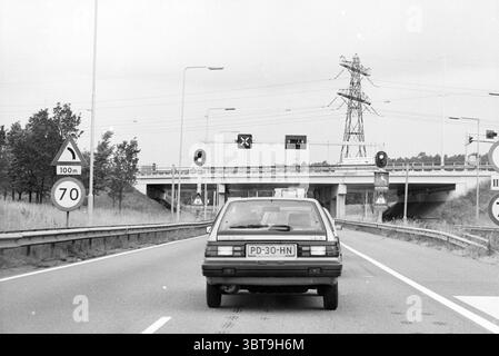 WESTERN Randweg in Richtung Velsertunnel Westelijke Randweg, Whizgle News, Dutch Desk, Niederlande, 1950 - 2000 am 06-1989. Das Bild enthält diese Themen. Die Szene zeigt eine breite, offene Straße, die sich bis in die Ferne erstreckt und von Grasland auf beiden Seiten flankiert wird. Ein Fahrzeug, das eher altmodisch wirkt, befindet sich im Vordergrund und fährt auf eine Reihe von Verkehrsschildern zu, die Straßenbedingungen und Geschwindigkeitsbegrenzungen anzeigen. Die Farbe des Autos ist gedämpft und fügt sich in den etwas bewölkten Himmel ein, der ein weiches, diffuses Licht über die gesamte Szene wirft. Im Hintergrund steht eine große Überführung mit Str. Stockfoto