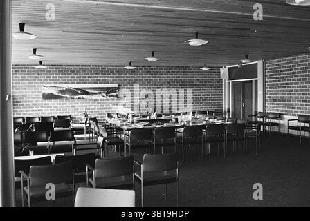 Ein Erholungsraum., Whizgle News, Dutch Desk, Niederlande, 1950 - 2000. Das Bild enthält diese Themen. Die Szene zeigt einen geräumigen Tagungsraum, der sich durch eine moderne, minimalistische Ästhetik auszeichnet. Die Wände bestehen aus hellen, strukturierten Ziegeln, die eine warme und dennoch industrielle Kulisse bieten. An einer Wand hängt ein großes, abstraktes Kunstwerk, das auf eine natürliche Landschaft mit fließenden Linien und weichen Formen hindeutet. In der Mitte des Raumes ist ein großer Tisch mit einheitlicher Anordnung angeordnet, der Teller, Gläser und Utensilien enthält, die auf eine Mahlzeit oder ein Treffen hinweisen. Die Stühle sind umrahmt Stockfoto