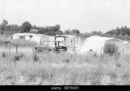 Verschiedene Strand- und Campingfotos IJmuiden und Spaarnwoude Camping Strand und Strände IJmuiden Niederlande, Whizgle News, Dutch Desk, Niederlande, 1950 - 2000 auf 24-07-1983. Dies sind die Themen im Bild. Die Szene zeigt einen rustikalen Campingplatz auf einem grasbewachsenen Feld, umgeben von Bäumen im Hintergrund. Verschiedene Zelte sind aufgehängt, einige scheinen als einfache Stoffstrukturen zu sein, während andere durch zusätzliche Abdeckungen robuster aussehen. Die Zelte sind locker angeordnet, was auf ein lässiges Layout hindeutet, und sie zeigen unterschiedliche weiß- und Beigetönen. Im Vordergrund, ein Jahrgang Stockfoto