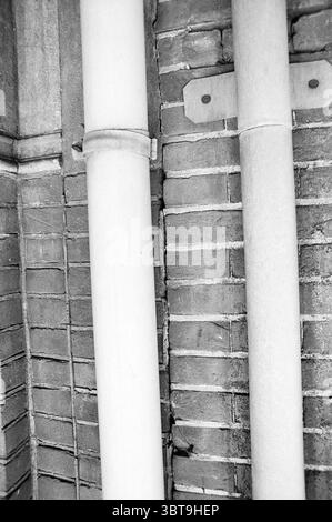 Drainpipe., Whizgle News, Dutch Desk, Niederlande, 1950 - 2000. Diese Themen werden in der Abbildung angezeigt. Die Szene zeigt eine Nahkomposition aus zwei vertikalen Rohren, die vor dem Hintergrund einer strukturierten Ziegelwand stehen. Die Rohre, gedämpft weiß lackiert, zeigen Anzeichen von Verschleiß, wobei einige Bereiche leicht angetaut oder verfärbt erscheinen. Ihre glatten Oberflächen stehen im Kontrast zur rauen, ungleichmäßigen Textur der roten Ziegelsteine, die verschiedene Tiefrot-Töne und subtile Orange- und Brauntöne aufweisen. Die Ziegelsteine sind in einem traditionellen Muster angelegt, was ein Gefühl von Struktur und Ordnung im roug schafft Stockfoto