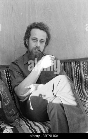 Porträt von Mr. Kater Porträts, Whizgle News, Dutch Desk, Niederlande, 1950 - 2000 am 06-12-1969. Diese Themen sind in der Abbildung dargestellt. In einem gemütlichen Raum mit weichem Tageslicht sitzt ein Mann bequem auf einer gemusterten Couch. Er hat lockige Haare und einen vollen Bart, was eine warme und ansprechbare Aura erzeugt. Er trägt ein schlichtes Hemd mit Knöpfen, das einen Kontrast zu den komplizierteren Textilmustern der Couch bildet. Hinter ihm befindet sich eine glatte Holzwand, die der Szene ein Gefühl von Wärme und Stabilität verleiht. Im Raum spielen schwache Schatten, die darauf hinweisen, dass die Tageszeit I ist Stockfoto