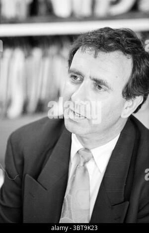 Herr Bonda Schiphol Schiphol, Whizgle News, Dutch Desk, Niederlande, 1950 - 2000 am 25-04-1993. Diese Themen werden in der Abbildung angezeigt. Die Szene zeigt einen Mann mit dunklem, leicht welligem Haar, der einen Moment der Interaktion in einem Gespräch festhält. Er trägt einen formellen schwarzen Anzug, ergänzt durch eine hellere Krawatte mit dezentem Muster, die ein professionelles Aussehen vermittelt. Der Hintergrund zeigt eine unscharfe Fülle von Dateien oder Dokumenten, die auf eine Büro- oder Arbeitsumgebung hinweisen. Die Farben sind gedämpft und monochromatisch, dominiert von Grau- und Schwarztönen, was die Ernsthaftigkeit von Th verstärkt Stockfoto