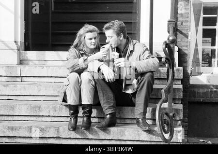 Junge Mann und Frau auf dem Gehweg der Hauptwache mit Eis ('schönes Wetter in Haarlem') Haarlem Grote Markt Niederlande, Whizgle News, Dutch Desk, Niederlande, 1950 - 2000 am 18-08-1988. Dies sind die Elemente im Bild. In dieser Szene sitzen zwei Individuen auf einer Steintreppe. Die Lage hat ein charmantes, urbanes Feeling, möglicherweise entlang eines Gehwegs in der Nähe eines Gebäudes mit großen Fenstern. Das Paar ist in einem ruhigen Moment verlobt, wobei eine Person einen Drink hält, während die andere sich etwas näher neigt und sich scheinbar intensiv auf den Drink konzentriert, wodurch ein Gefühl von Intimität entsteht. Sie sind Bot Stockfoto