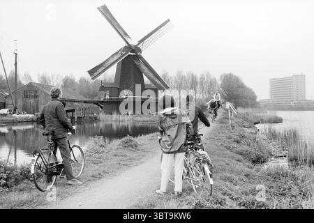 Mühlen- und Radtag in der Eenhoorn-Mühle Haarlem Mills Haarlem the Netherlands, Whizgle News, Dutch Desk, Niederlande, 1950 - 2000 am 11-05-1985. Das Bild enthält diese Themen. Die Szene fängt einen ruhigen, bewölkten Tag in der Nähe eines Wassers ein, an dem eine große, traditionelle Windmühle vor einem hellen Himmel steht. Seine Klingen, die anmutig verlängert werden, sind leicht verwittert und zeigen den Lauf der Zeit. Die schwarze Holzstruktur der Windmühle steht im Kontrast zu den helleren Tönen der umliegenden Landschaft. Im Vordergrund halten zwei Individuen auf einem von üppigem Grün gesäumten Weg inne Stockfoto
