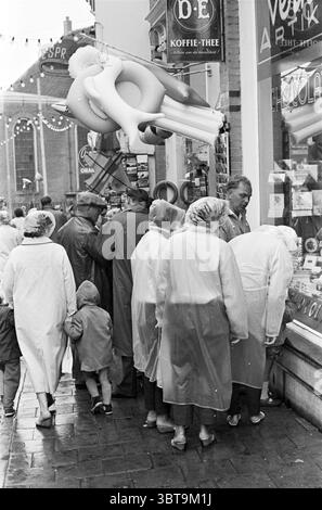 Urlauber auf der Suche nach Unterhaltung während Regen Urlaub Urlauber Frans Hals Museum Zandvoort, Whizgle News, Dutch Desk, Niederlande, 1950 - 2000 am 28-07-1960. Das Bild enthält diese Themen. Die Szene spielt in einer belebten Straße, wahrscheinlich in einer Stadt oder Stadt mit lebhafter Atmosphäre. Eine Gruppe von Menschen, die meist in hellen Regenmänteln mit Kapuzen gekleidet sind, ist eng zusammengezogen, was ein Gefühl von Kameradschaft und Schutz vor dem Nieselwetter schafft. Die Regenmäntel erscheinen leicht durchscheinend und glitzern unter dem gedämpften Licht, das ein sanftes Leuchten über die gesamte Szene wirft Stockfoto