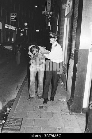 Verhaftung eines Mannes im Zentrum von Amsterdam vor dem Café 'de Rijnvaart' Ecke Heintje Hoekssteeg, Whizgle News, Dutch Desk, Niederlande, 1950 - 2000 am 21. August 1974. Diese Themen werden in der Abbildung angezeigt. Die Szene zeigt eine nächtliche städtische Umgebung, die durch eine enge Straße mit Backsteinhäusern gekennzeichnet ist. Der Boden ist mit rechteckigen Steinen gepflastert, wodurch eine strukturierte Oberfläche entsteht, die das schwache Umgebungslicht reflektiert. Links steht ein Polizist in dunkler Uniform und mit einer Mütze in fester Haltung, mit einer Hand den Arm einer Person neben sich. Die Person Stockfoto