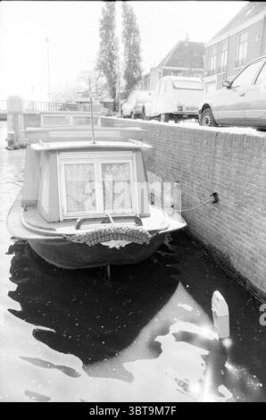 Vergnügungsboot im Kanal., Whizgle News, Dutch Desk, Niederlande, 1950 - 2000. Diese Themen sind in der Abbildung dargestellt. Die Szene zeigt einen ruhigen Kanal, der von Ziegelmauern gesäumt ist, wo ein kleines Boot sanft auf dem Wasser schwimmt. Das Boot ist in tiefen Rot- und Weißtönen gestrichen und hat ein charmantes, leicht rustikales Aussehen, das durch einen Spitzenvorhang hervorgehoben wird, der durch das Fenster sichtbar ist. Um das Boot herum reflektiert das Wasser ein weiches, gedämpftes Licht, das ein Gefühl der Ruhe schafft. Die beschauliche Oberfläche des Kanals steht im Kontrast zu der strukturierteren Ziegelwand, die als Grenze dient und Brauntöne und zeigt Stockfoto