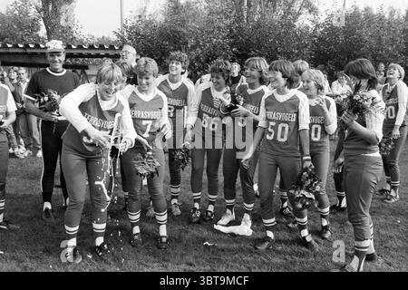 Terrasvogels - HCAW Softball, Whizgle News, Dutch Desk, Niederlande, 1950 - 2000 am 12-09-1982. Dies sind die Elemente im Bild. Im Vordergrund feiert eine Gruppe junger Athleten, wahrscheinlich im Teenageralter oder Anfang 20, auf einem grasbewachsenen Feld. Sie tragen koordinierte Sportuniformen, vor allem in leuchtenden Rot- und Weißtönen mit Zahlen auf den Trikots. Die Uniformen werden durch passende Shorts und sportliche Socken ergänzt. Einige der Menschen springen vor Begeisterung, während andere lachen und Momente der Freude teilen. Ein paar halten Blumen hoch oder klein Stockfoto