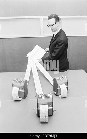 Mann mit drei Fahrkartenautomaten (Transportunternehmen?), Whizgle News, Dutch Desk, Niederlande, 1950 - 2000 am 11-1967. Die Abbildung zeigt diese Themen. Die Szene zeigt einen Mann, der an einem Tisch steht und an einer Aktivität beteiligt ist, an der mehrere Maschinen beteiligt sind. Er trägt einen formellen schwarzen Anzug mit hellem Hemd und Krawatte, was einen Sinn für Professionalität verkörpert. Die Haltung des Mannes deutet auf Konzentration hin, während er ein Dokument hält und möglicherweise die Informationen überprüft. In der Mitte des Tisches befinden sich drei Geräte, die jeweils mechanischen Rechnern oder Datenverarbeitungsmaschinen ähneln. Sie haben eine Retro-Aesth Stockfoto