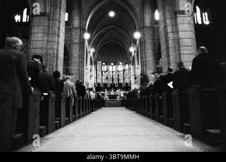 Innenraum der St. Bavo Kathedrale während der Messe Haarlem Westergracht Niederlande., Whizgle News, Dutch Desk, Niederlande, 1950 - 2000. Dies sind die Elemente im Bild. Die Szene entfaltet sich in einem großen gewölbten Raum, der ein Gefühl von Ehrfurcht und Hochmut ausstrahlt. Majestätisch erheben sich aufragende Säulen, die eine hohe Decke mit komplizierten architektonischen Details tragen. Das Licht, das durch Buntglasfenster filtert, wirft ein Kaleidoskop gedämpfter Farben auf den Steinboden, was die spirituelle Atmosphäre unterstreicht. Reihen von Holzbänken erstrecken sich den Mittelgang hinunter, gefüllt mit Menschen, die stehen Stockfoto