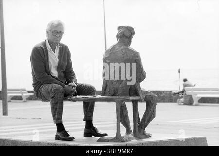 Kees Verkade bei Statuen Zandvoort Heemstede Verkade Kees Bildhauer Heemstede Niederlande, Whizgle News, Dutch Desk, Niederlande, 1950 - 2000 am 18-06-1987. Dies sind die Themen im Bild. In dieser Szene sitzt ein älterer Mann auf einer Bank und wirkt kontemplativ. Er trägt eine Brille und eine Strickjacke über einem leichten Hemd und eine karierte Hose, die seiner Kleidung einen klassischen Touch verleiht. Er hat einen nachdenklichen Ausdruck im Gesicht, blickt auf den Betrachter oder verliert sich vielleicht in Reflexion. Neben ihm sitzt eine Bronzestatue eines anderen Mannes auf derselben Bank. Die Statue hat ein verwittertes Aussehen, Showcas Stockfoto