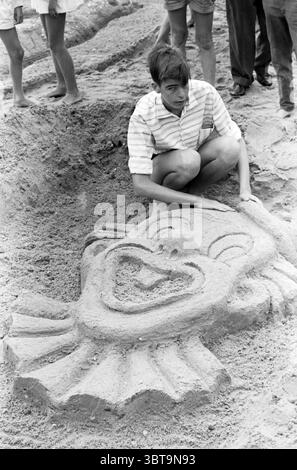 Sandbauwettbewerb in Zandvoort Pipo the Clown von Sieger André Morren aus Amsterdam. Sandskulpturen Sandburgen. Zandvoort, Whizgle News, Dutch Desk, Niederlande, 1950 - 2000 am 17-08-1960. Dies sind die Elemente im Bild. In einer lebhaften Strandszene konzentriert sich ein junger Mensch intensiv auf die Gestaltung einer großen Sandkreation. Die Figur mit gestreiftem Hemd und Shorts kniet neben einer detaillierten Sandskulptur, die einem verspielten Clowngesicht ähnelt, mit übertriebenen Details und einem liebenswürdigen Lächeln. Die Textur des Sandes ist fein granuliert, sodass klare Konturen und Entschärfung möglich sind Stockfoto