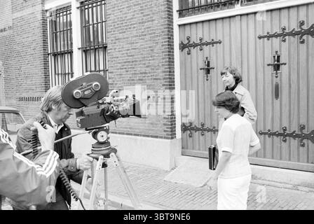TV. Gefängnisaufzeichnungen Fernsehen, Whizgle News, Dutch Desk, Niederlande, 1950 - 2000 am 24-06-1977. Diese Themen werden in der Abbildung angezeigt. In einer geschäftigen städtischen Umgebung erleben zwei Personen eine Szene, die von einer großen, verzierten Holztür mit komplizierten schwarzen Schmiedearbeiten eingerahmt wird. Die satte braune Farbe der Tür steht im Kontrast zur Backsteinfassade des Gebäudes, die eine Mischung aus tiefem Rot und gedämpftem Braun aufweist und der Kulisse eine historische und erdige Atmosphäre verleiht. Eine Person, die näher am Betrachter positioniert ist, bedient eine Vintage-Filmkamera, die auf einem stabilen Stativ montiert ist. Diese Kamera, mit ihrer mechanischen Stockfoto