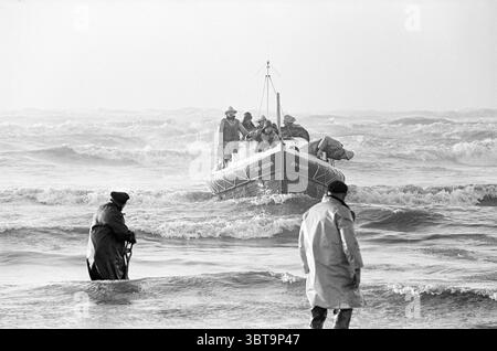 Rettung auf altmodische Weise am Strand von IJmuiden North South Hollandse Vervoersmaatschappij N.V. NZ, Whizgle News, Dutch Desk, Niederlande, 1950 - 2000 am 14-01-1965. Dies sind die Themen im Bild. In einer dynamischen Meereslandschaft krachen und Rollen Wellen mit einem schäumenden weißen Wappen, was auf eine energiegeladene und turbulente Atmosphäre hindeutet. Der Horizont ist in ein sanftes, gedämpftes Grau gehüllt und verschmilzt nahtlos mit einem nebeligen Himmel darüber, was der Szene eine etwas düstere und melancholische Stimmung verleiht. Im Vordergrund steht eine Einzelfigur auf dem nassen Sand, der teilweise in Wasser getaucht ist. In einem dunklen, schweren coa Stockfoto