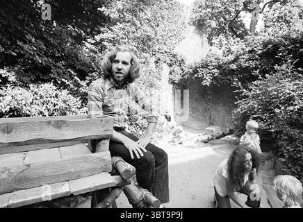 Wim Schalken Jugendberater Persons, Whizgle News, Dutch Desk, Niederlande, 1950 - 2000 am 20-08-1978. Das Bild enthält diese Themen. Die Szene fängt eine ruhige Umgebung im Freien ein, geprägt von üppigem Grün und einer reichen Palette von Blättern, die die Komposition einfangen. Eine rustikale Holzbank aus Baumstämmen nimmt den Vordergrund ein, wo ein Mann mit lockigen Haaren ruhig sitzt. Seine Kleidung, ein kariertes Hemd, fügt sich in die natürliche Kulisse ein und trägt zur entspannten Atmosphäre bei. Im Hintergrund ist das Grün dick und lebhaft, was eine Einladung zur Natur andeutet. Sonnenlicht filtert durch die l Stockfoto