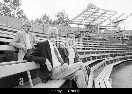 Stadtrat Wilma und Mitarbeiter im Stadion 1986 Baseball-Weltmeisterschaften, Whizgle News, Dutch Desk, Niederlande, 1950 - 2000 am 12.06.1986. Das Bild enthält diese Themen. In einer geräumigen, offenen Umgebung sitzen drei Personen bequem auf einer Reihe angeordneter Bänke, die Teil eines größeren erhöhten Gebäudes sind. Die Sitzmöbel bestehen aus hellem Metall und Holzlatten, was eine minimalistische Ästhetik schafft. Der Hintergrund zeigt Reihen leerer Sitze, die in die Ferne fallen, was darauf hindeutet, dass der Veranstaltungsort für Veranstaltungen, möglicherweise Sport oder Aufführungen konzipiert ist. Auf der einen Seite gibt es Bäume casti Stockfoto