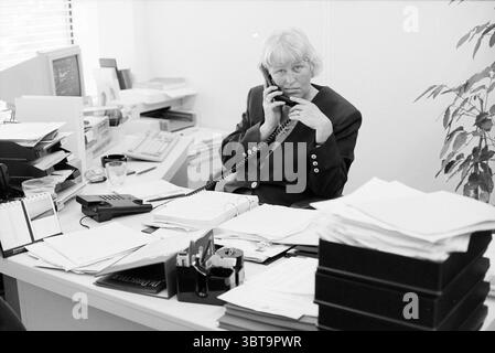 Mrs. Broers kam Haarlem Haarlem the Netherlands, Whizgle News, Dutch Desk, Niederlande, 1950 - 2000 am 24-05-1993. Diese Themen werden in der Abbildung angezeigt. Die Szene zeigt eine Frau, die an einem überdachten Schreibtisch in einer Büroumgebung sitzt. Sie hat ein kurzes, helles Haar und trägt einen dunklen Blazer, der einen Kontrast zu den helleren Tönen ihrer Umgebung bildet. Ihr Ausdruck vermittelt ein Gefühl der Konzentration und vielleicht eine leichte Frustration, während sie am Telefon spricht, wobei ihr Kopf leicht zur Seite geneigt ist. Der Schreibtisch ist voller Papierkram, was auf eine geschäftige Arbeitsumgebung hinweist. Verschiedene Dokumente A Stockfoto