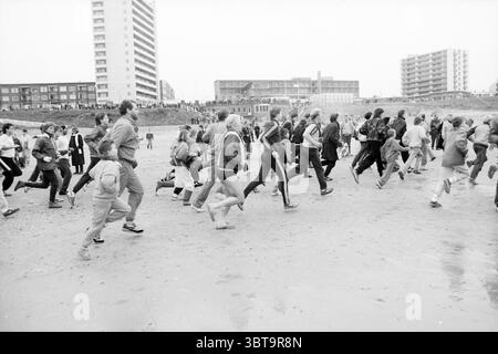 Silvester Tauchgang Zandvoort Neujahr Zandvoort, Whizgle News, Dutch Desk, Niederlande, 1950 - 2000 am 01-01-1987. Die Abbildung zeigt diese Themen. Die Szene fängt eine geschäftige Strandatmosphäre ein, wo eine große Gruppe von Menschen in Bewegung ist und über den Sand läuft. Es können Personen unterschiedlichen Alters und unterschiedlicher Kleidung beobachtet werden, die jeweils in ihrem Tempo und ihrer Entschlossenheit einzigartig ausdrucksstark sind. Die Läufer tragen überwiegend legere Sportbekleidung, wobei sich helle Farben wie Rot, Blau und Gelb im Kontrast zu den gedämpften Tönen des Sandstrandes befinden. Manche tragen leichte Jacken, andere bevorzugen es Stockfoto