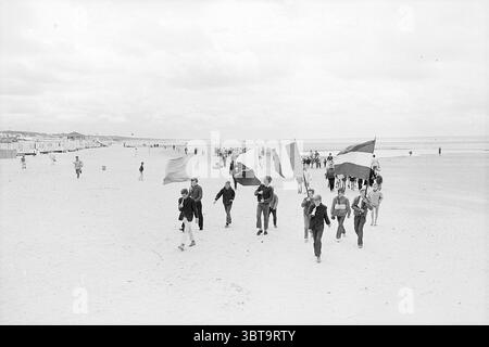 Abfahrtsgruppe vom Strand zum Zirkus Abfahrt, Whizgle News, Dutch Desk, Niederlande, 1950 - 2000 am 25-07-1968. Diese Themen sind in der Abbildung dargestellt. Eine vielfältige Gruppe von Einzelpersonen spaziert entlang eines breiten Sandstrandes, deren Fußstapfen kleine Eindrücke im hellen Sand erzeugen. Die Szene ist voll von einer Mischung aus Aktivität und Freizeit, während andere Menschen im Hintergrund spazieren gehen, sich treffen und an verschiedenen Strandaktivitäten teilnehmen. Die Gruppe im Vordergrund hält farbenfrohe Fahnen, jede winkt in der sanften Brise. Die Flaggen zeigen eine Vielzahl von Farben, die einen lebhaften Kontrast zum Mu erzeugen Stockfoto