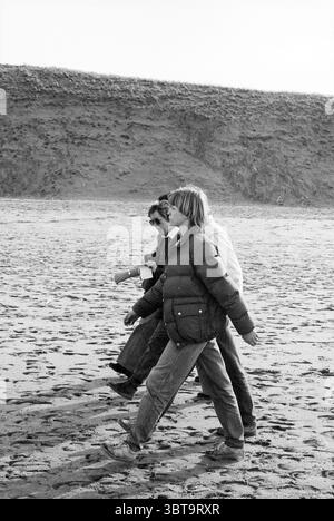 Winter Walk HD Haarlems Dagblad etc. Und IJmuider Courant und Koeri Walking, Whizgle News, Dutch Desk, Niederlande, 1950 - 2000 am 14.02.1982. Die Abbildung zeigt diese Themen. Die Szene zeigt drei Personen, die an einem Sandstrand entlang laufen, wobei ihre Bewegungen synchronisiert sind, während sie gezielt voranschreiten. Jede Person trägt legere Kleidung, die für einen kühlen Tag am Ufer geeignet ist. Die erste Figur links hat dunkles Haar, trägt eine dunkle Jacke über einem helleren Hemd und einer Jeans. Neben ihnen trägt ein zweites Individuum eine geschwollene Winterjacke, die wahrscheinlich Wärme gegen die kühle Brise bietet, mit ihrem hai Stockfoto